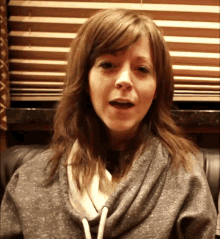 a woman in a grey sweatshirt is sitting in front of blinds