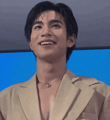 a man wearing a tan jacket and a necklace smiles