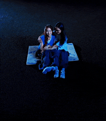 two women are sitting on a blue blanket on the ground at night