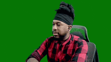 a man in a red and black plaid shirt is sitting in a chair with a green screen behind him