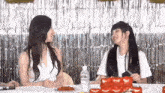 two girls are sitting at a table with a cake and a bottle of water .