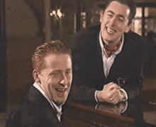 two men in suits and bow ties are laughing at a piano
