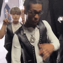 a man wearing glasses and a vest with studs is standing next to another man