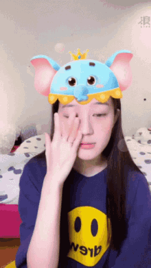 a girl wearing a blue shirt with a yellow smiley face and a blue elephant hat