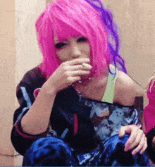 a woman with pink and purple hair is sitting down and covering her mouth with her hands .
