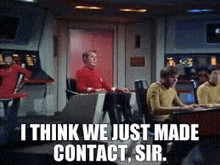 a group of men are sitting at a desk in a room and talking to each other .