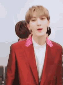 a young man wearing a red suit jacket and a pink collar is standing in front of a group of people .