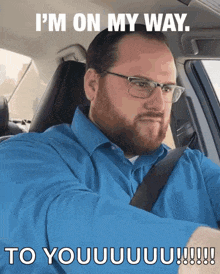 a man wearing glasses and a blue shirt is driving a car and says i 'm on my way to you