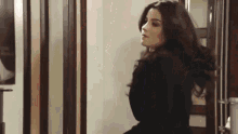 a woman in a black jacket is standing in front of a door with her hair blowing in the wind .