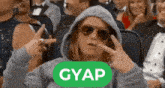 a woman wearing a hoodie and sunglasses is sitting in a crowd with a green sign that says gyap on it