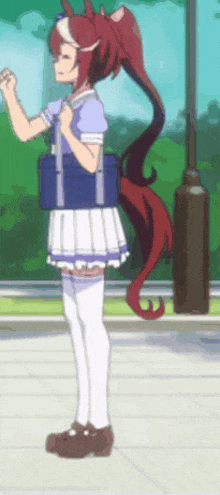 a girl with red hair and a horse tail is standing on a sidewalk holding a blue bag .