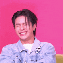 a young man wearing a blue denim jacket is laughing with his eyes closed .