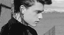 a black and white photo of a young man leaning against a fence looking at the water .