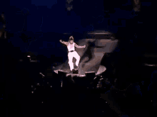 a man in a white tank top and white skirt is dancing on a stage with his arms outstretched .