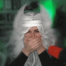 a person wearing a white wig with horns is covering their mouth with their hands