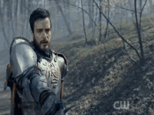 a man in armor is standing in the middle of a forest with a cw logo behind him