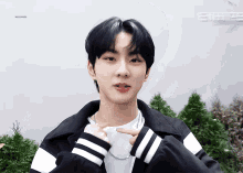 a young man wearing a white t-shirt and a black and white jacket is pointing at something