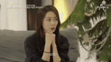 a woman is sitting on a couch with her hands folded and a channel logo in the background .