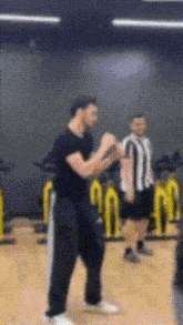 a man in a black shirt is standing in a gym with a referee behind him .