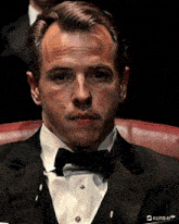 a man in a tuxedo sits in a red chair with kling ai written on the bottom of his shirt