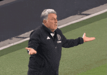 a man wearing an adidas jacket stands on a soccer field with his arms outstretched