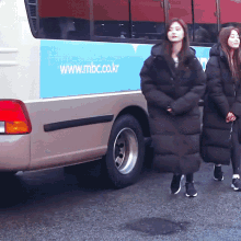 two women standing in front of a bus that says www.mbc.co.kr on the side