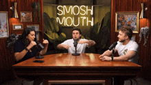 three people sit at a table in front of a smosh mouth neon sign