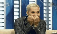 a man with blonde hair is covering his mouth with his hands while sitting in front of a wall with the letter g on it