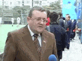 a man in a suit and tie is standing in front of a soccer field talking into a microphone ..