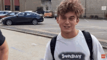 a young man wearing a sunday t-shirt smiles in a parking lot
