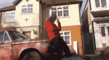 a man in a red hoodie is sitting on the back of a rusty car in front of a house .