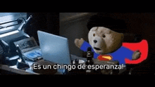 a teddy bear dressed as superman is sitting in front of a laptop .
