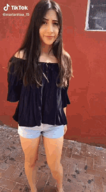 a woman in a black off the shoulder top and shorts stands in front of a red wall