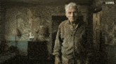an older man in a dirty sweater stands in a room