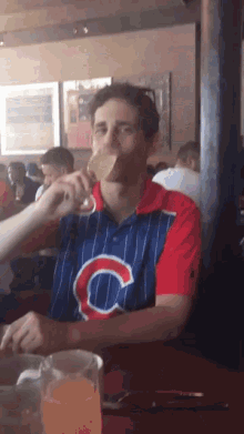 a man wearing a blue and red shirt with the letter c on it drinks from a glass
