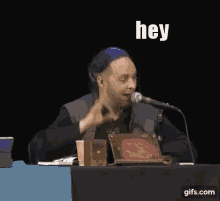 a man is sitting at a table talking into a microphone and saying `` hey cool '' .