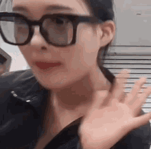 a woman wearing glasses and a black jacket is waving at the camera .