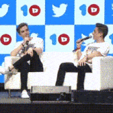 two men are sitting in chairs on a stage with a microphone in front of a twitter background .