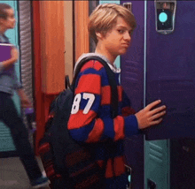 a young boy wearing a striped sweater with the number 87 on it is standing in front of a purple locker .