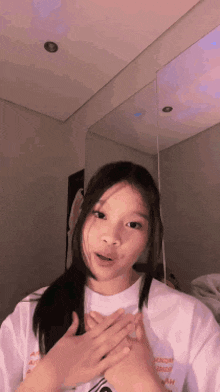 a young girl in a white t-shirt is standing in front of a mirror holding her hands to her chest .