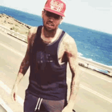 a man wearing a tank top and a red hat stands on the side of the road near the ocean