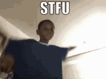 a boy in a blue shirt is standing in a room with the words stfu on the ceiling .