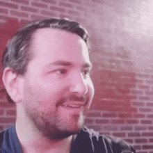 a man with a beard smiles in front of a brick wall
