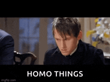 a man in a suit is sitting at a table with a sign that says homo things behind him .