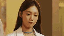 a woman wearing a white coat and a blue necklace is standing in a hallway .