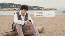 a man sits on a log on the beach looking at his phone with a message coming out of his phone that says ca va mieux