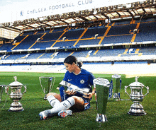 a woman sits on the grass holding a trophy in front of the chelsea football club