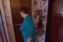 a woman in a blue shirt is standing in front of a door with a floral wallpaper .