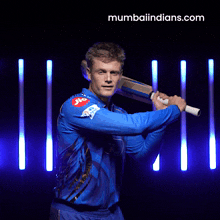 a man in a blue shirt is holding a bat with the website mumbaiindians.com behind him