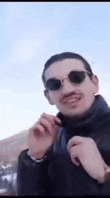 a man wearing sunglasses and a mustache is standing in front of a blue sky and smiling .
